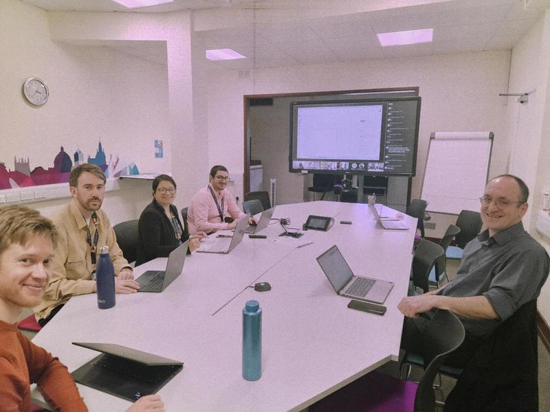 Image of 5 team members sitting around a meeting room table, with laptops and smiling at the camera